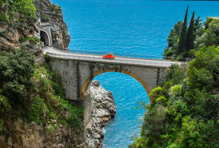 A car navigates an arch bridge spanning a seaside gorge.