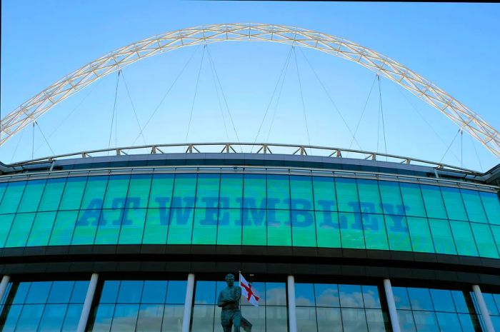 A vast array of glass windows adorns the upper section of a grand stadium, with a massive metal arch towering overhead.