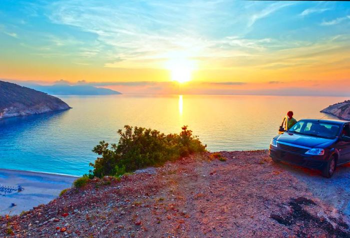 A woman leaning against an open car door parked on the beach, with a stunning sunset casting colors over the sea.