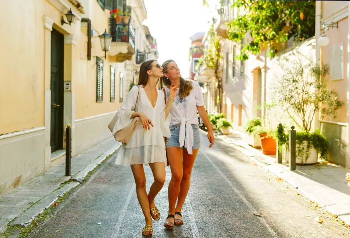 Two women exploring a sunlit alleyway flanked by vibrant houses.