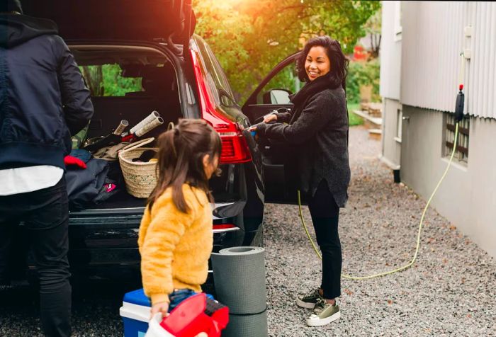 A woman recharges an electric vehicle while a man and a young girl organize items in the trunk.