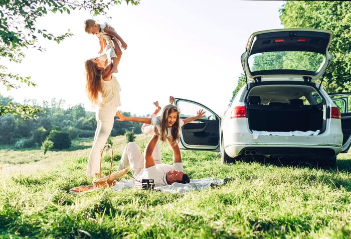 A woman stands while lifting a boy into the air next to a man lying on a blanket, who is raising a girl beside a white car.