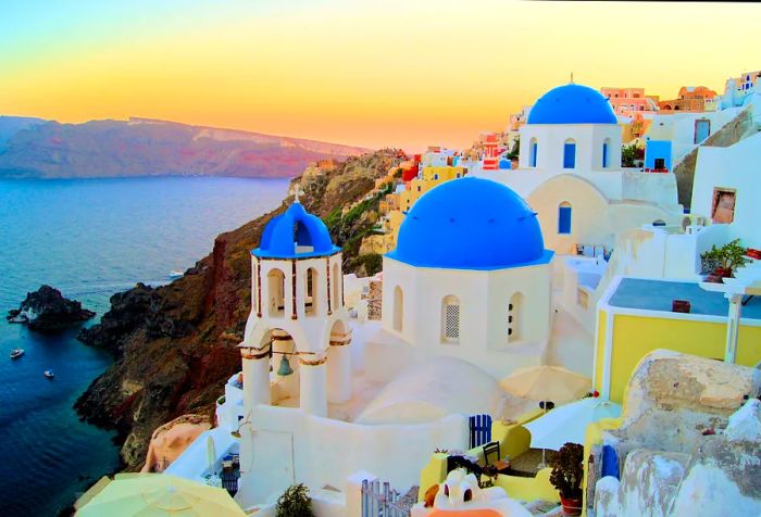 A picturesque blue dome church with a belfry perched on a cliffside village under a glowing orange sky.