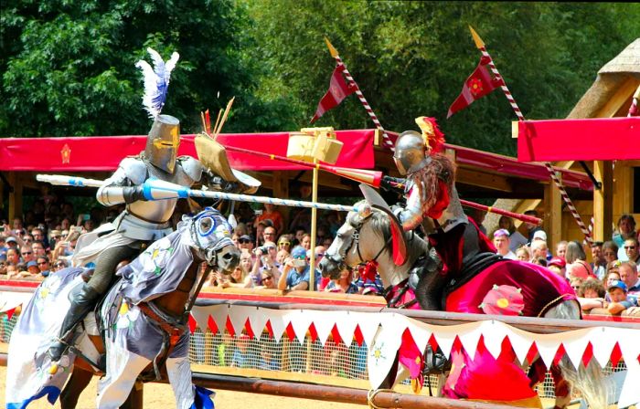 Two armored knights on horseback face each other, brandishing jousting sticks in an exciting battle reenactment with an audience of spectators.