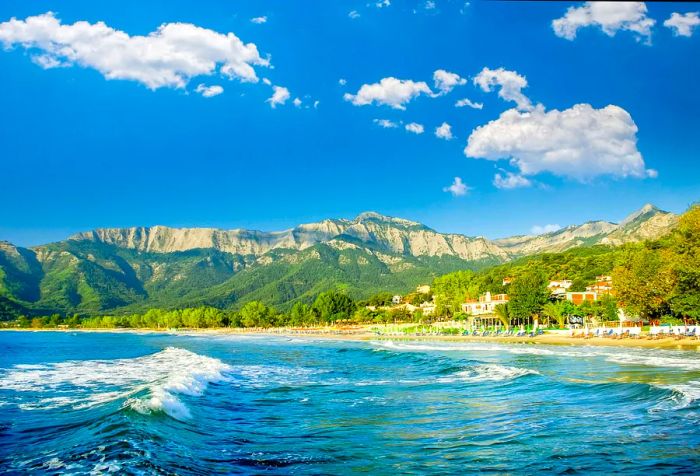 Waves crash against the shore, with lush trees and a breathtaking mountain range as the backdrop.