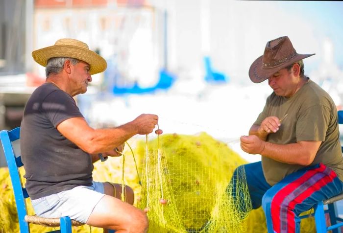 Two men in hats relax on blue wooden chairs, busily mending yellow fishing nets.