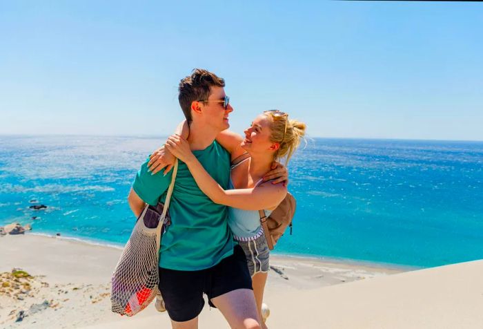 A young couple stands atop the sand dunes at the beach, gazing into each other's eyes, with the man carrying a net shoulder bag and the woman in a spaghetti-strap blouse, creating a romantic beach vibe.