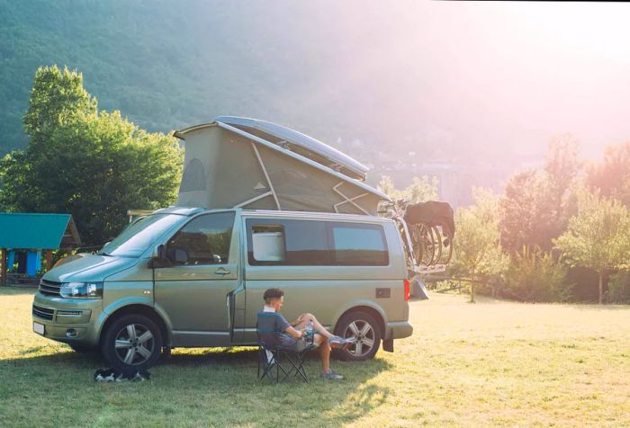 A person relaxes on a folding chair beside a sleeping dog, next to a grey camper van fitted with a bike rack and a rooftop tent.