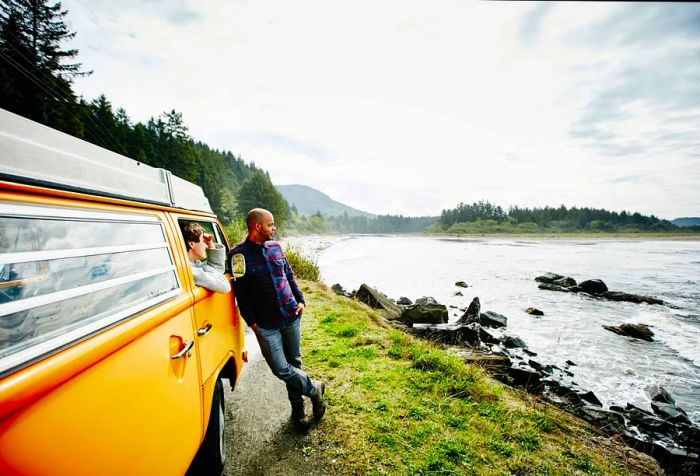 A woman looking out the window while a man leans against the side of a van, gazing at the ocean.