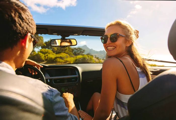 A cheerful young woman enjoys the ride in a convertible as a man drives them along the road.