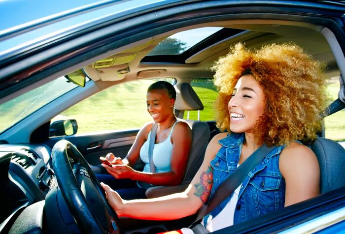 A woman with curly blonde hair drives a car with a friend seated beside her in the passenger seat.