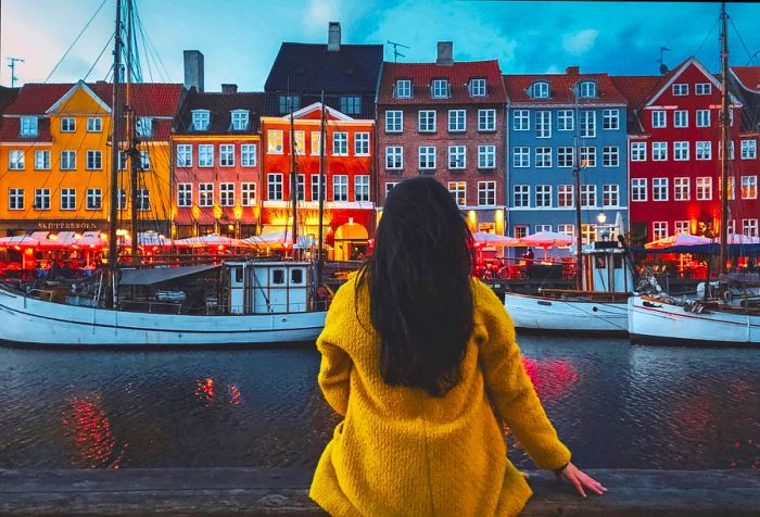 A person in a yellow cardigan sits on a wooden railing, looking out over the harbor and the boats docked nearby.