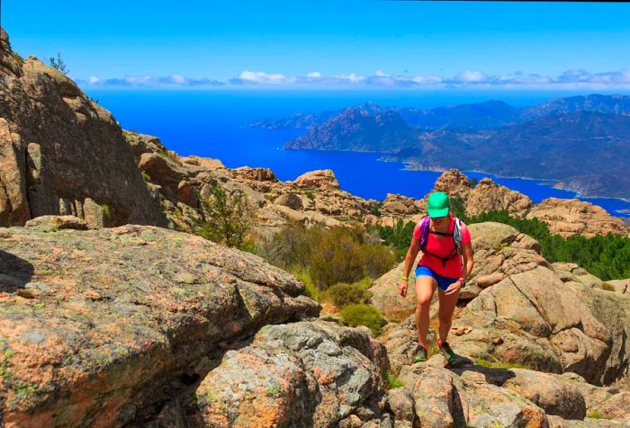 A woman in a pink shirt and hat navigates a rugged mountain trail, overlooking the valleys and ocean in the distance.