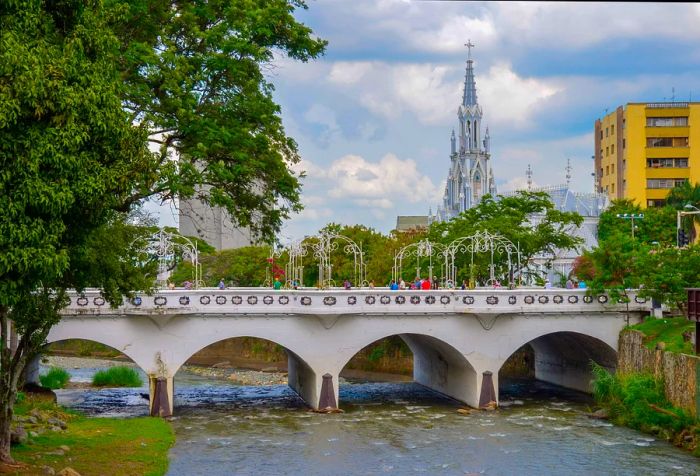 Pedestrians traverse a white arch bridge over a serene river, framed by the striking silhouette of a church steeple in the background.