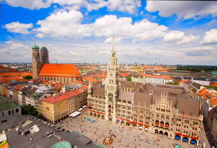 A bustling public square in front of a gothic building featuring a clock tower, with a church boasting twin spires in the background.