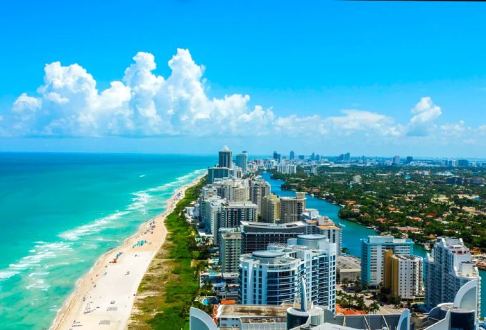 A row of towering buildings stretches along a serene river beside the vibrant turquoise sea on a bright, sunny day.