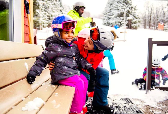 A happy mother and child dressed in colorful winter gear, enjoying the scenery next to skiers in the background.