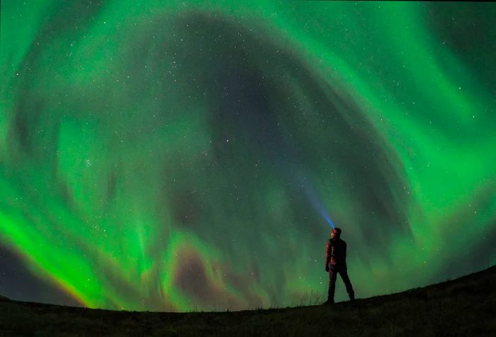 A man stands in the foreground with a lit headlamp, captivated by the dazzling Northern Lights overhead.