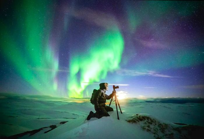 A photographer captures the stunning Northern Lights illuminating the sky, kneeling beside his tripod to get the perfect shot.