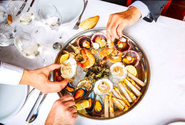 A group of hands sharing delicious finger foods, featuring mussels, clams, and oysters from a stainless steel tray.