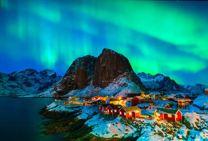 The green Northern Lights swirl across the sky above a quaint fishing village, with cabins lining the rocky, snow-covered coastline.