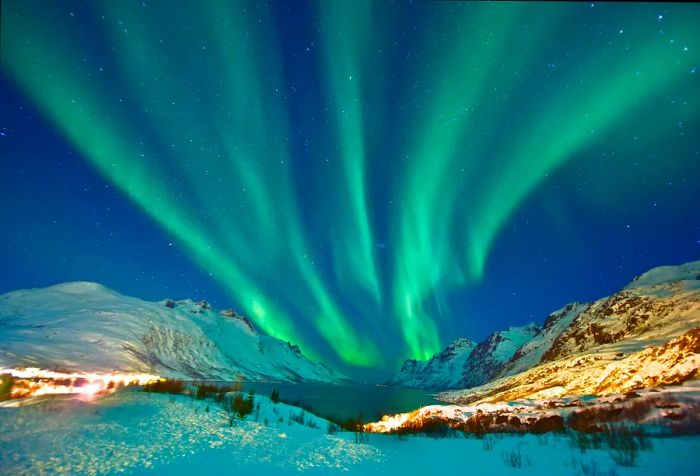 The breathtaking Aurora Borealis twirls in the night sky above a snow-clad mountain range, casting vibrant reflections on a tranquil, frozen lake below.