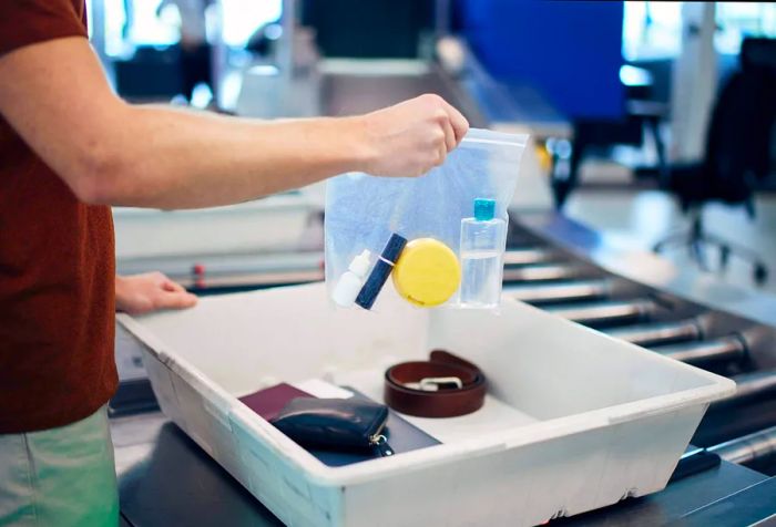 Airport security check prior to departure. A traveler holds a plastic bag containing liquids above a laptop and personal belongings.