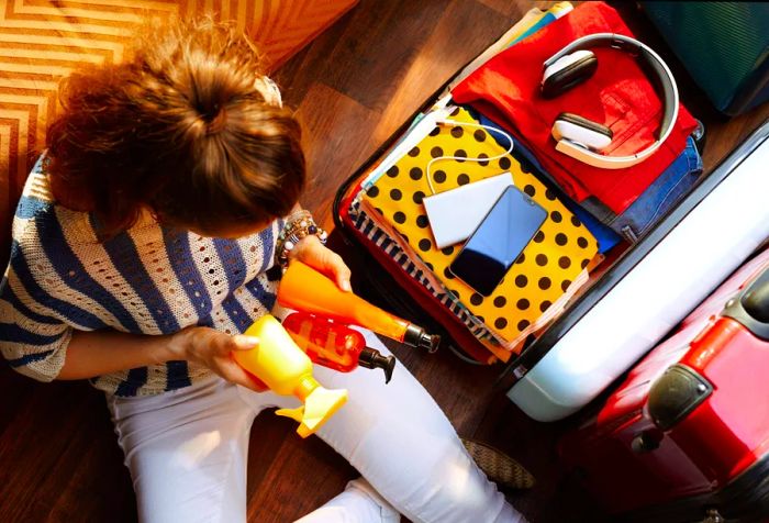 Overhead view of a modern woman in white pants and a striped blouse, packing summer cosmetics into an open suitcase on a sunny day.