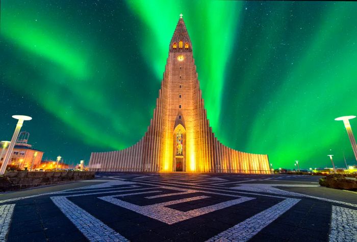 A nighttime view of Hallgrímskirkja, a church featuring curved spires and wings beneath the green northern lights in a starry sky.