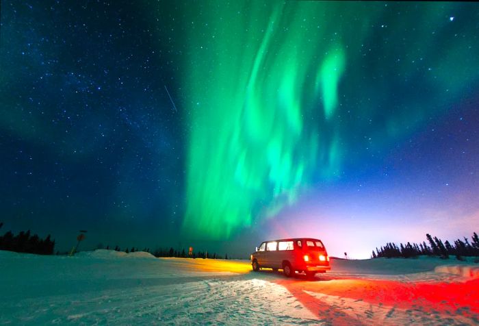 A van sits in a snowy landscape beneath the vibrant northern lights and sparkling stars.