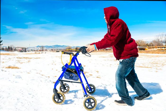 A side view of a young, disabled Hispanic Millennial man using a walker for assistance while navigating a snow-covered park sidewalk in Denver, Colorado, on a winter day.