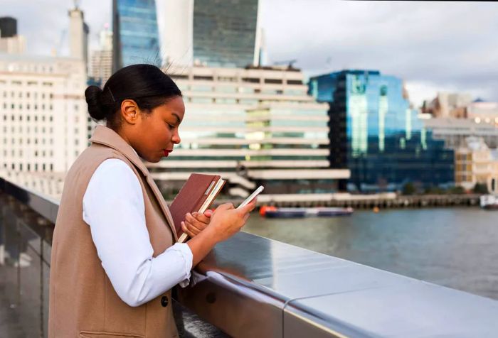 A woman stands on a bridge, engrossed in a book while using her smartphone.