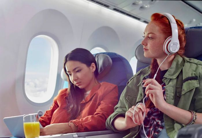 Two women on a flight: one engrossed in her laptop while the other enjoys music through headphones.