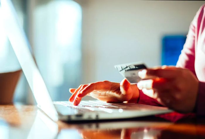 The right hand is busy typing on a laptop, while the left hand holds a credit card.