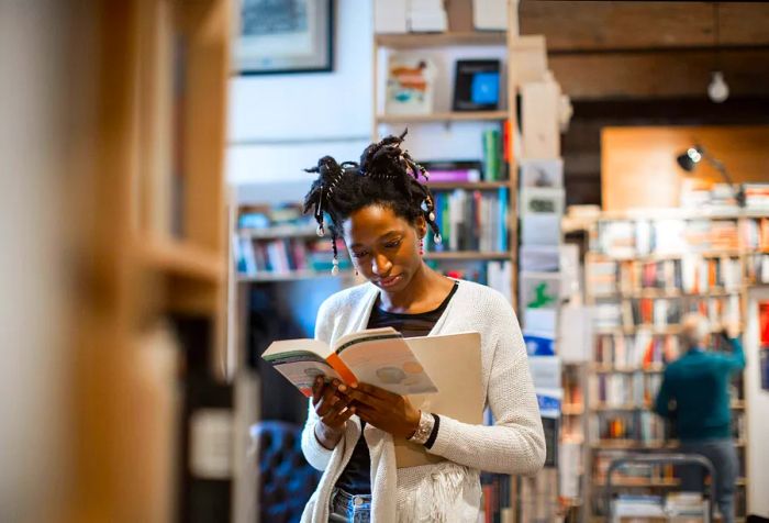 A young woman is absorbed in a book while surrounded by the bustling shelves of a lively bookstore.