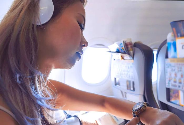 A woman wearing headphones glances at her watch while seated on an airplane.