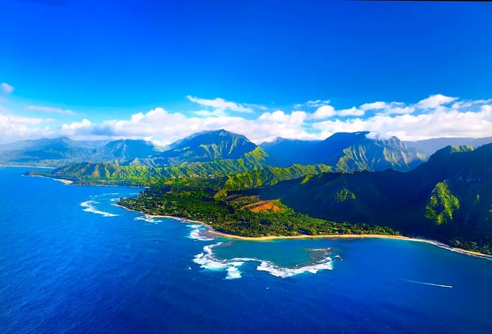 A serene blue sea with gentle waves and a mountain range draped in lush greenery against a partly cloudy sky.