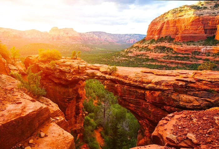 A striking sandstone arch rises above a forested area, offering views of colorful, layered rock mountains.