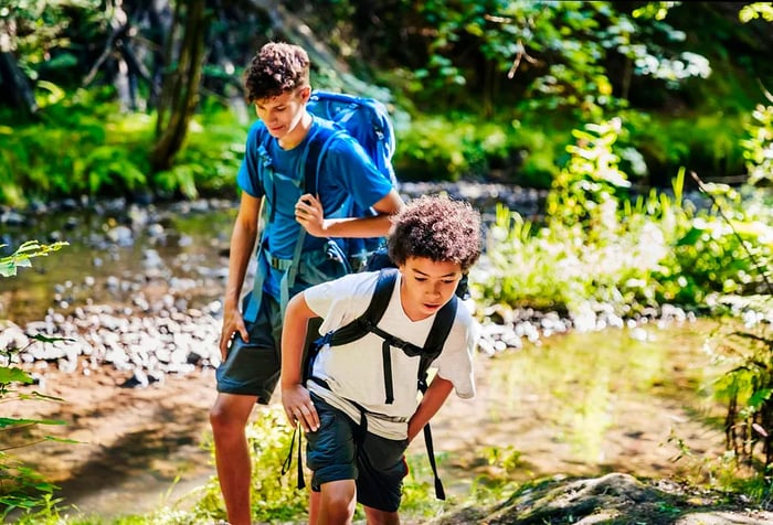 Two boys with backpacks navigate a river trail through the forest.