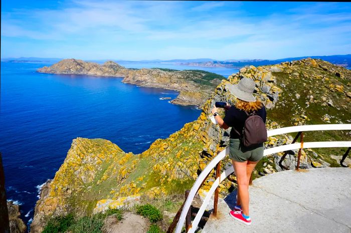 A woman captures a photo from a viewpoint, showcasing the rugged island that juts into the ocean below.