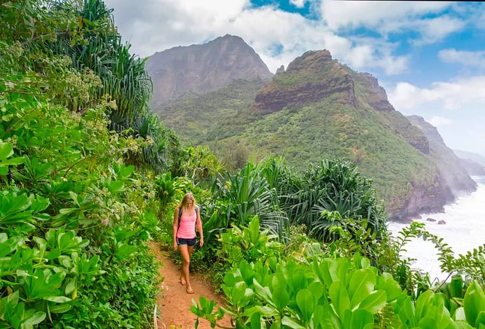 A woman hikes barefoot along a hillside trail in a forest, with stunning ocean views crashing against the distant mountainous coastline.