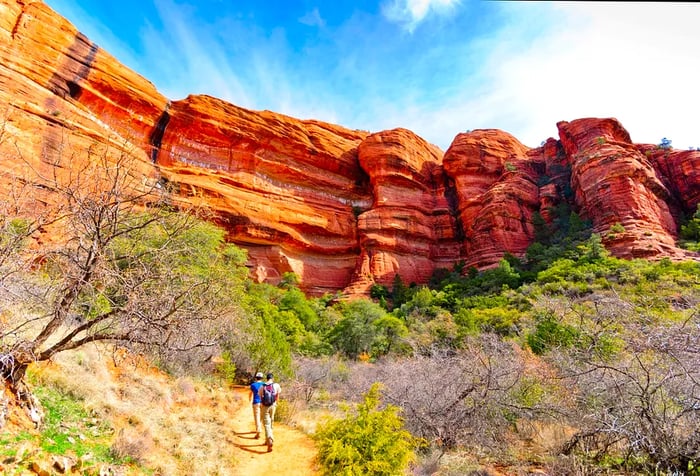 Two male friends stroll along a dirt path, surrounded by dry, lush trees as they approach the breathtaking red rock formations.