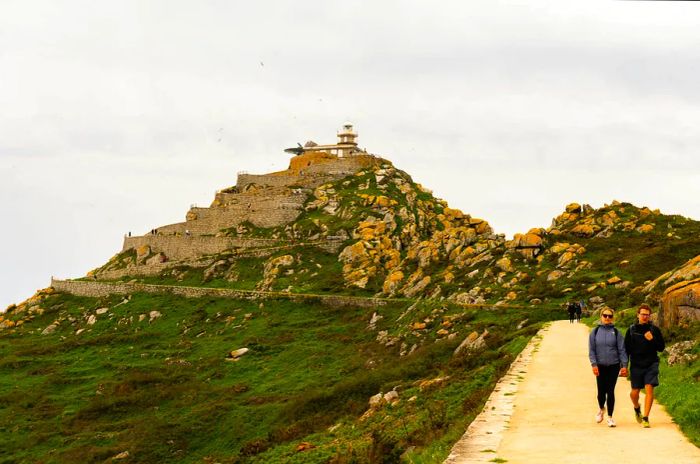 Two individuals stroll along a path on the island, making their way from a clifftop lighthouse.