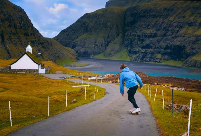 A man in a light blue hoodie skateboards down a winding road toward a white church, with a serene lake at the foot of rugged mountains.