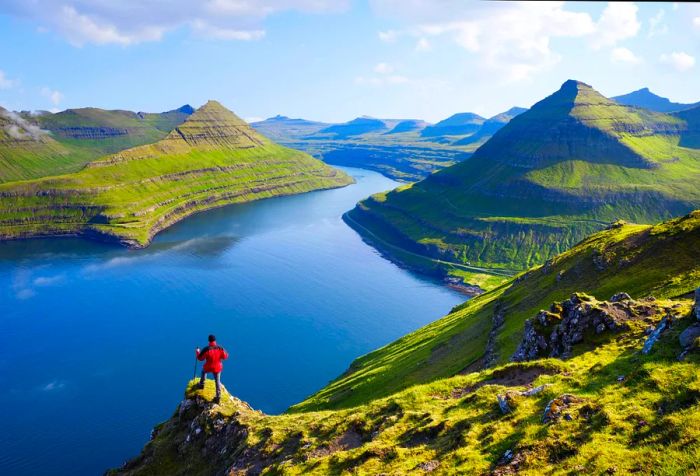 A figure in a red jacket stands on a narrow cliff, gazing at the tranquil lake nestled among the rocky mountains of an island.