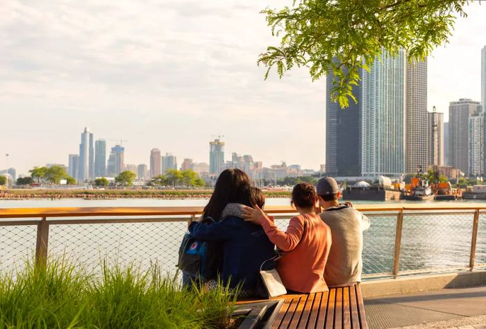 Groups of people unwind in a lush, waterfront park with towering skyscrapers as a backdrop.