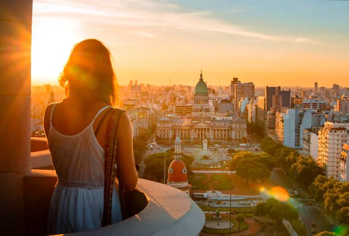 A person gazes from a balcony, enjoying a sweeping view of the expansive city that cradles a tranquil park below, revealing a dynamic urban landscape.