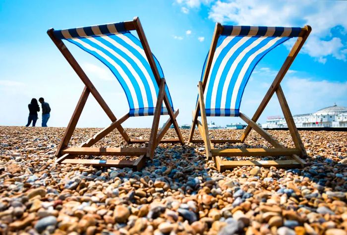 Two deck chairs sit on a pebbly beach, with a silhouette of a couple in the distance.