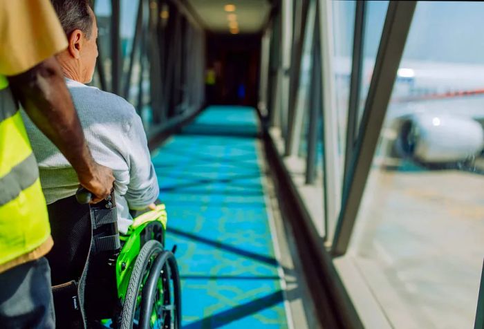 An airport staff member assists a man in a wheelchair as they navigate the boarding bridge.