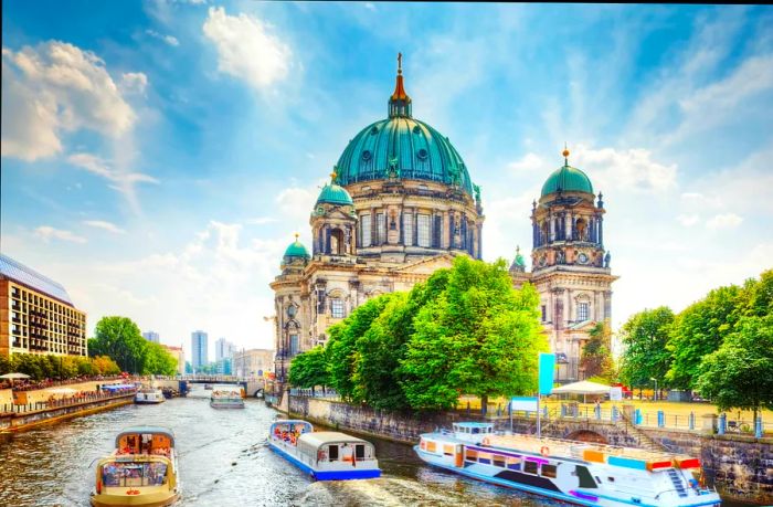 Passenger boats glide along the river Spree on a sunny day, with the Berliner Dom (Berlin Cathedral) in the background.